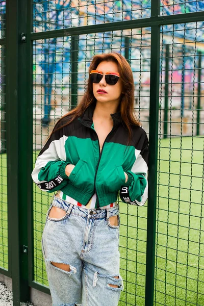 Portrait of a girl clothes in the style of the 90s sports style, jacket, jeans bananas, sunglasses, sports field residential area