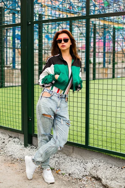 Portrait of a girl clothes in the style of the 90s sports style, jacket, jeans bananas, sunglasses, sports field residential area