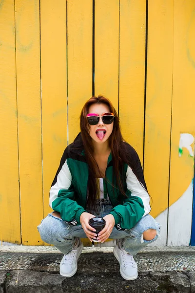 Portrait of a girl in clothes in the style of the 90s, sporty style, jacket, jeans, bananas, sunglasses. Lady on the background of wooden colored background, drinking morning coffee