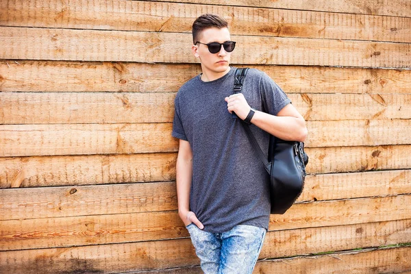 Hipster style guy. handsome guy with glasses and a fashionable hairdo. vintage photo. The guy standing near the old wooden wall. In the hands of a fashion backpack and a lion, sunglasses