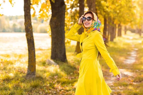 Stylish beautiful lady in a yellow raincoat and head scarf, sunglasses, autumn walk in the woods, fashion trend of the season