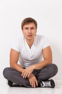 Beyaz tişörtlü ve gri pantolonlu yakışıklı adam beyaz arka planda oturuyor.