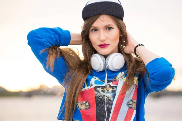 Stylish teenage girl with long hair, headphones and a cap is posing near a river