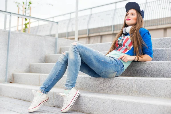 一个戴着耳机 牛仔裤坐在台阶上的时髦女孩 — 图库照片