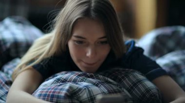 Odada rahat bir yatakta dinlenirken akıllı telefon kullanan genç bir kadın.