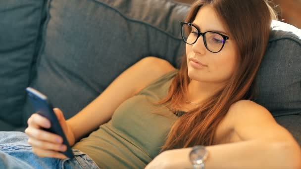 部屋の中で居心地の良いソファでリラックスしながらスマートフォンを使用して若い女性 — ストック動画