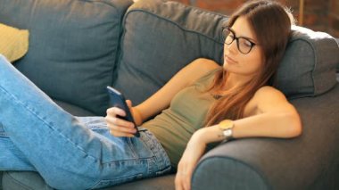 Genç bir kadın odasındaki rahat koltukta dinlenirken akıllı telefon kullanıyor.