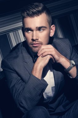 Çizgili bir arka planda spot ışıkları altında oturan takım elbiseli yakışıklı bir adamın portresi.