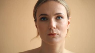 Sarı saçlı, bej arka planda kameraya poz veren doğal bir genç kadın portresi. 