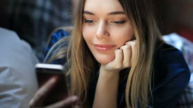 Genç bir kadın odasındaki rahat koltukta dinlenirken akıllı telefon kullanıyor.