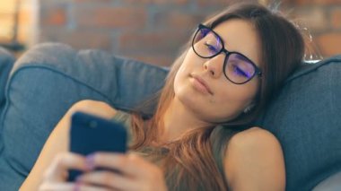 Genç bir kadın odasındaki rahat koltukta dinlenirken akıllı telefon kullanıyor.