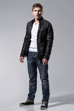 Deri ceketli, kot pantolonlu ve beyaz tişörtlü yakışıklı bir adam gri arka planda poz veriyor.