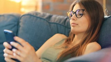 Genç bir kadın odasındaki rahat koltukta dinlenirken akıllı telefon kullanıyor.