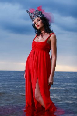 Sahilde poz veren, taç ve kırmızı elbiseli zarif bir kadın.