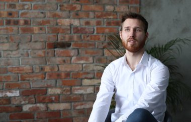 Beyaz gömlekli yakışıklı genç adam tuğla duvar arkasında poz veriyor.