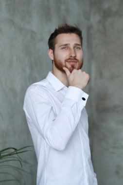 Beyaz gömlekli yakışıklı adam gri duvarda poz veriyor.