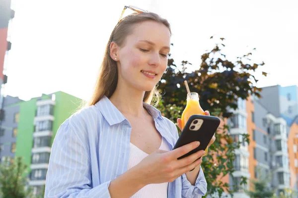 Young smiling woman drinks natural orange juice from a paper tube and looks at the phone on the background of buildings, bottom view. The concept of advertising juices and takeaway drinks