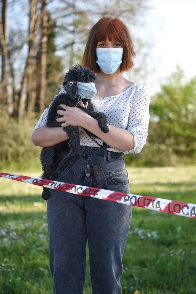 Girl with surgical mask face protection with a puppy in her arms,  behind a red and white striped ribbon. Nature background. COVID19