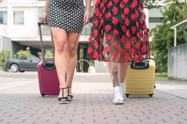 Şehrin çiçekli caddesinde bavullarıyla yürüyen iki zarif turist kadının kesilmiş fotoğrafı..