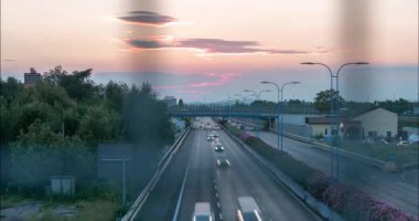 Otobanın günbatımında yol güvenlik tellerinin arkasındaki üstgeçitten 4K 'lık bir video zaman atlaması. Ön planda tel örgü olan trafik akışı.