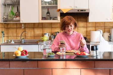 Genç, tatlı bir kadın mutfağında yemek yiyor ve akıllı telefon kullanıyor..