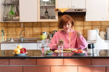 Genç, tatlı bir kadın mutfağında yemek yiyor ve akıllı telefon kullanıyor..