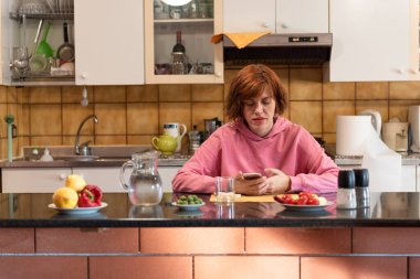 Genç, tatlı bir kadın mutfağında yemek yiyor ve akıllı telefon kullanıyor..