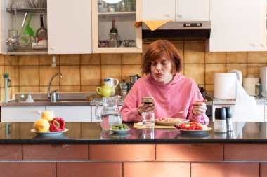 Genç, tatlı bir kadın mutfağında yemek yiyor ve akıllı telefon kullanıyor..