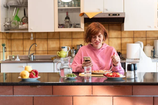 Genç, tatlı bir kadın mutfağında yemek yiyor ve akıllı telefon kullanıyor..