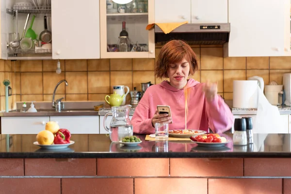 Genç, tatlı bir kadın mutfağında yemek yiyor ve akıllı telefon kullanıyor..