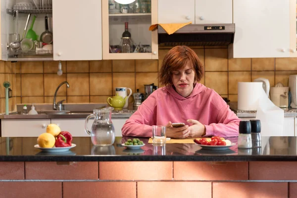 Genç, tatlı bir kadın mutfağında yemek yiyor ve akıllı telefon kullanıyor..