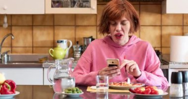 Genç tatlı kız mutfağında yemek yiyor ve cep telefonu kullanıyor. Kızıl saçlı kız..