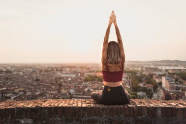 Gün batımında güneşi tutan yoga pozisyonunda olan sadık bir kadının arka ışığının önünde ya da sıcak bir arka planı olan güzel bir şehirde güneşin doğuşunda.