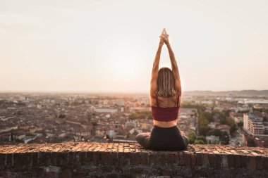 Gün batımında güneşi tutan yoga pozisyonunda olan sadık bir kadının arka ışığının önünde ya da sıcak bir arka planı olan güzel bir şehirde güneşin doğuşunda.