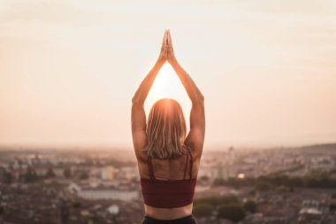 Gün batımında güneşi tutan yoga pozisyonunda olan sadık bir kadının arka ışığının önünde ya da sıcak bir arka planı olan güzel bir şehirde güneşin doğuşunda.