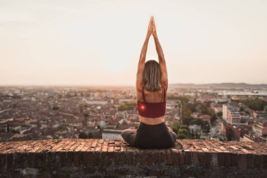 Gün batımında güneşi tutan yoga pozisyonunda olan sadık bir kadının arka ışığının önünde ya da sıcak bir arka planı olan güzel bir şehirde güneşin doğuşunda.