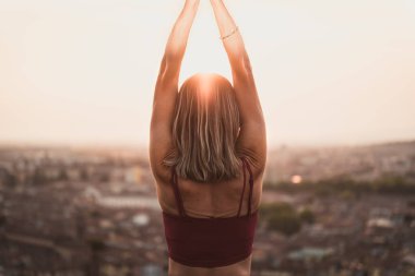 Gün batımında güneşi tutan yoga pozisyonunda olan sadık bir kadının arka ışığının önünde ya da sıcak bir arka planı olan güzel bir şehirde güneşin doğuşunda.