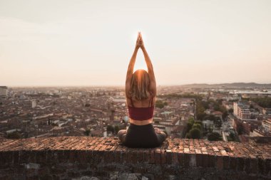 Gün batımında güneşi tutan yoga pozisyonunda olan sadık bir kadının arka ışığının önünde ya da sıcak bir arka planı olan güzel bir şehirde güneşin doğuşunda.