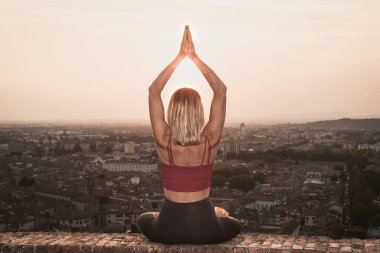 Gün batımında güneşi tutan yoga pozisyonunda olan sadık bir kadının arka ışığının önünde ya da sıcak bir arka planı olan güzel bir şehirde güneşin doğuşunda.