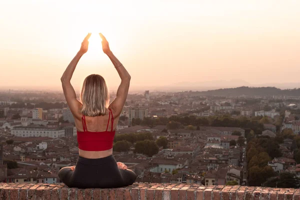 Gün batımında güneşi tutan yoga pozisyonunda olan sadık bir kadının arka ışığının önünde ya da sıcak bir arka planı olan güzel bir şehirde güneşin doğuşunda.