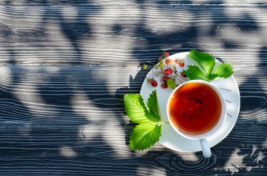 Koyu ahşap bir masada yabani olgun çilekli bir fincan çay. Terasta güneşli bir günde parlak gölgeler.