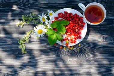 Tahta bir masanın üzerinde yabani olgun çilekler ve bir fincan çay. Terasta güneşli bir günde parlak gölgeler.