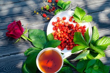 Tahta bir masanın üzerinde yabani olgun çilekler ve bir fincan çay. Terasta güneşli bir günde parlak gölgeler.
