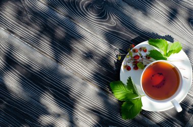 Koyu ahşap bir masada yabani olgun çilekli bir fincan çay. Terasta güneşli bir günde parlak gölgeler.