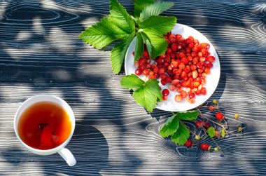 Tahta bir masanın üzerinde yabani olgun çilekler ve bir fincan çay. Terasta güneşli bir günde parlak gölgeler.