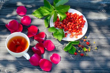 Tahta bir masanın üzerinde yabani olgun çilekler ve bir fincan çay. Terasta güneşli bir günde parlak gölgeler.