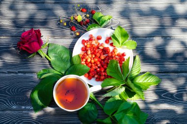 Tahta bir masanın üzerinde yabani olgun çilekler ve bir fincan çay. Terasta güneşli bir günde parlak gölgeler.