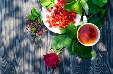 Tahta bir masanın üzerinde yabani olgun çilekler ve bir fincan çay. Terasta güneşli bir günde parlak gölgeler.