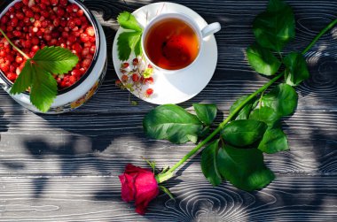 Tahta bir masanın üzerinde yabani olgun çilekler ve bir fincan çay. Terasta güneşli bir günde parlak gölgeler.