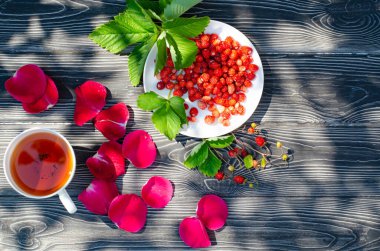 Tahta bir masanın üzerinde yabani olgun çilekler ve bir fincan çay. Terasta güneşli bir günde parlak gölgeler.